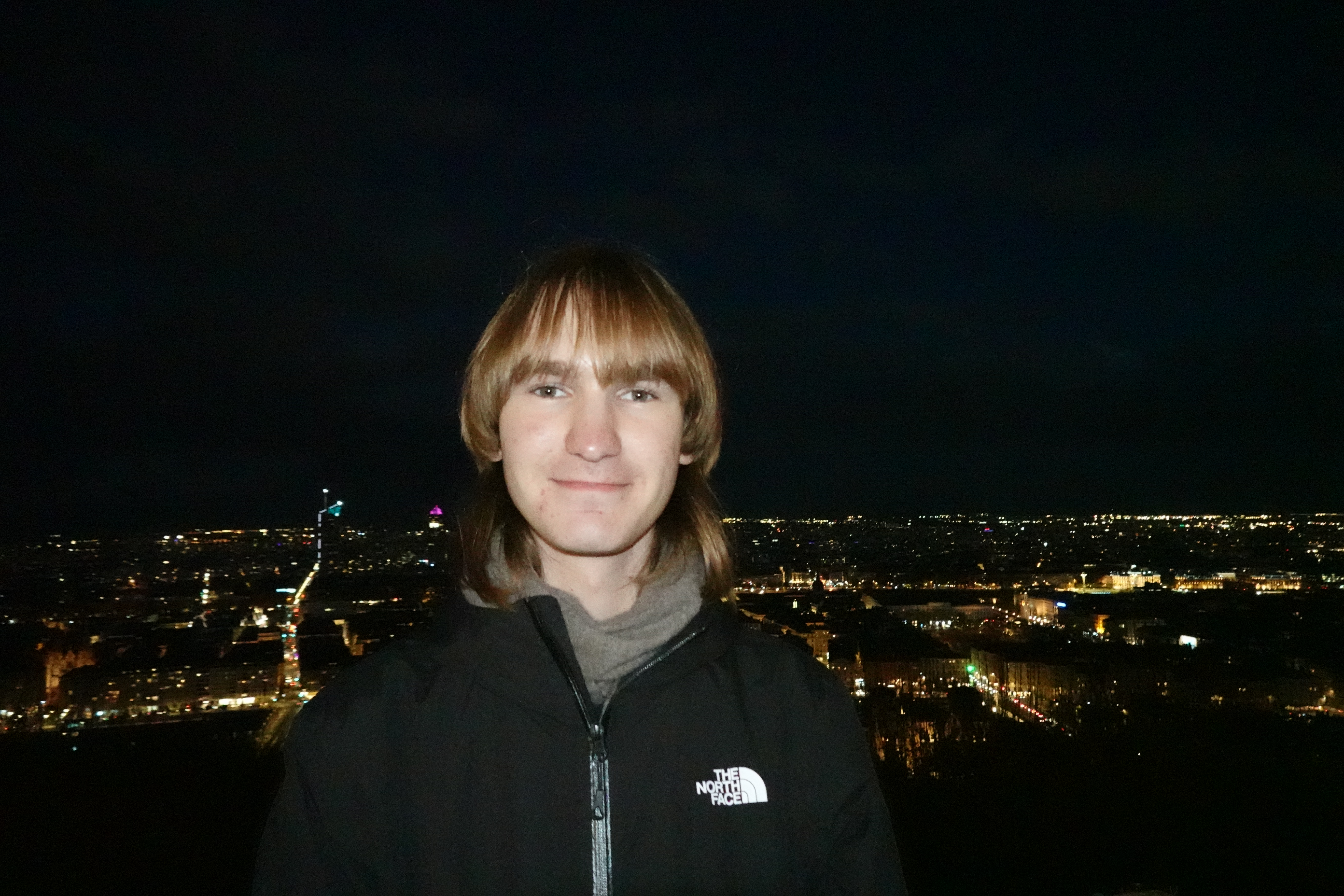 A flash photo of me standing and smiling, overlooking the evening lights of Lyon.